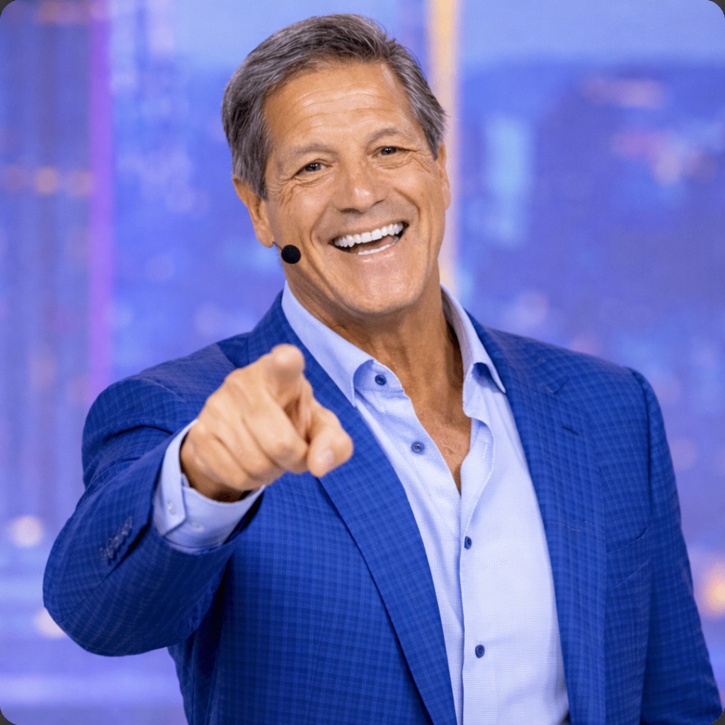 John Assaraf smiling during a business coaching session, wearing professional attire in a studio-style setting